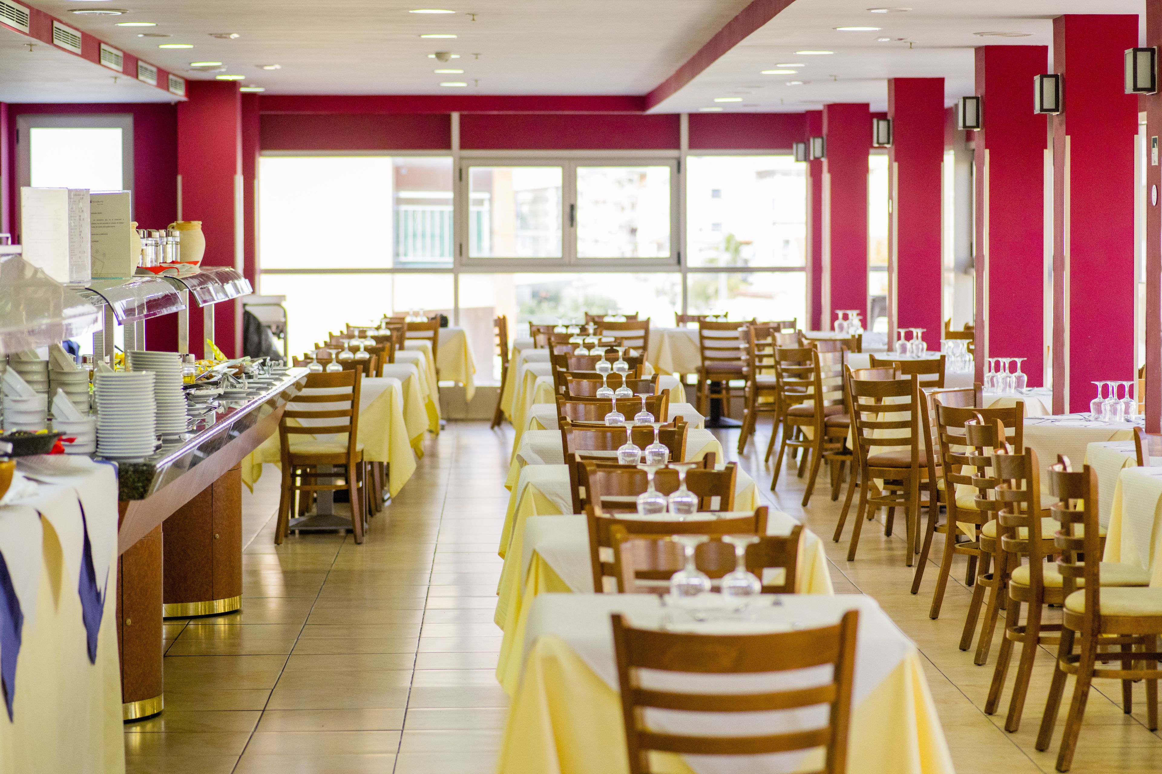 Zona pranzo con diversi tavoli e sedie, un'area buffet con varie pietanze e ampie finestre che lasciano entrare la luce naturale.