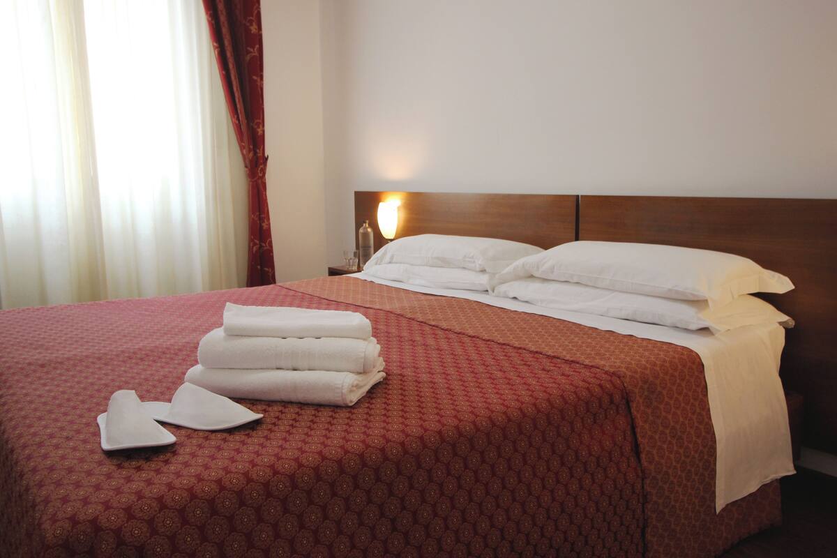 A hotel room with a neatly made bed, a bedside table with a lamp, and a window with curtains.