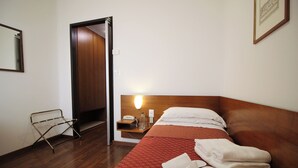A hotel room with a wooden floor, a bed with a red patterned bedspread, a nightstand with a lamp, a mirror, and a door leading to another room.