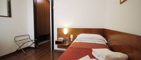 A hotel room with a wooden floor, a bed with a red patterned bedspread, a nightstand with a lamp, a mirror, and a door leading to another room.