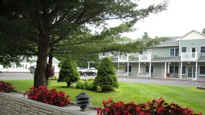 Un edificio con fachada verde, un porche cubierto y un jardín delantero con flores rojas y arbustos podados.