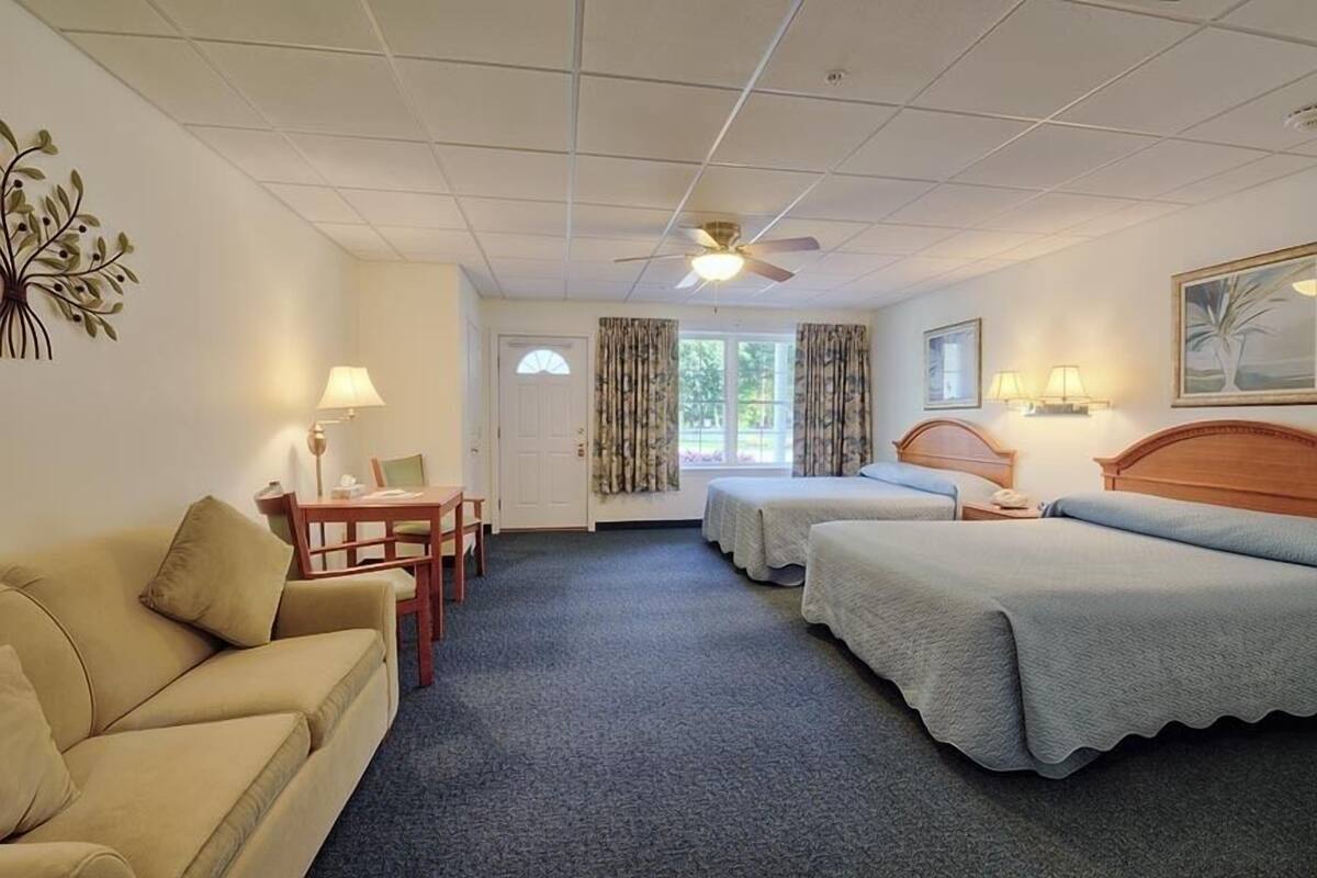Et hotelværelse med en sofa, to enkeltsenge, et skrivebord med stol, en loftsvifte og en dør med et vindue.