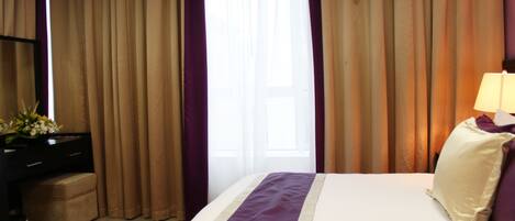 A hotel room with a bed, curtains, a desk with a lamp, and a small table with flowers.