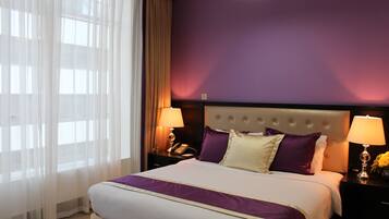 A hotel room with a large bed, two bedside tables with lamps, a nightstand with a phone, and sheer curtains on the window.