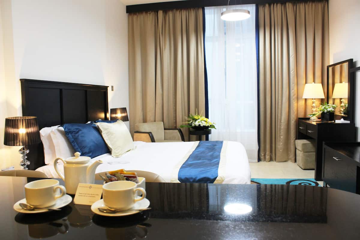 Une chambre d’hôtel comprenant un lit, une table de chevet, des lampes, un bureau avec une chaise et une fenêtre avec des rideaux.