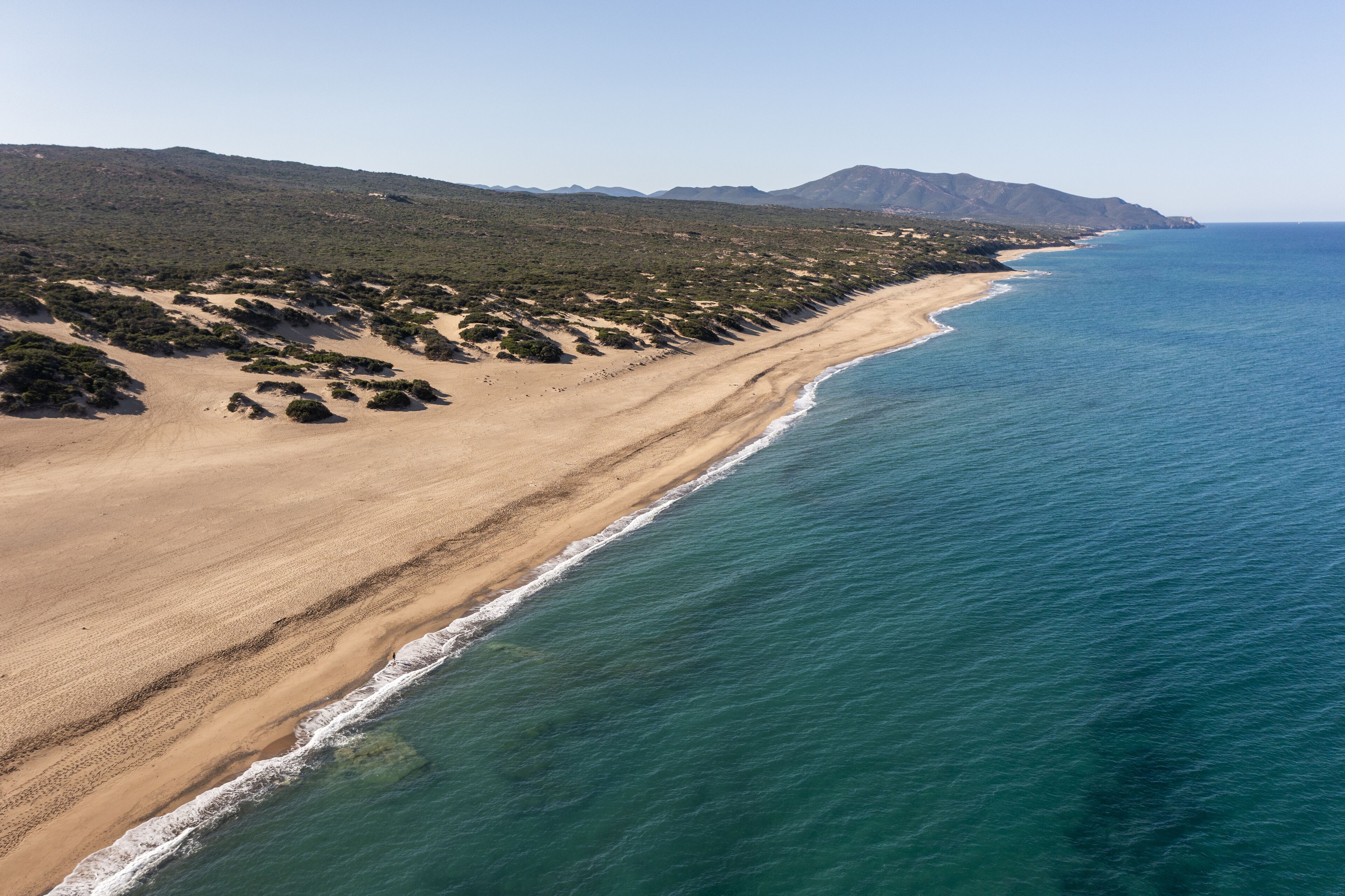 An aerial view of a coastal landscape with a sandy beach, green vegetation, and a clear blue ocean.