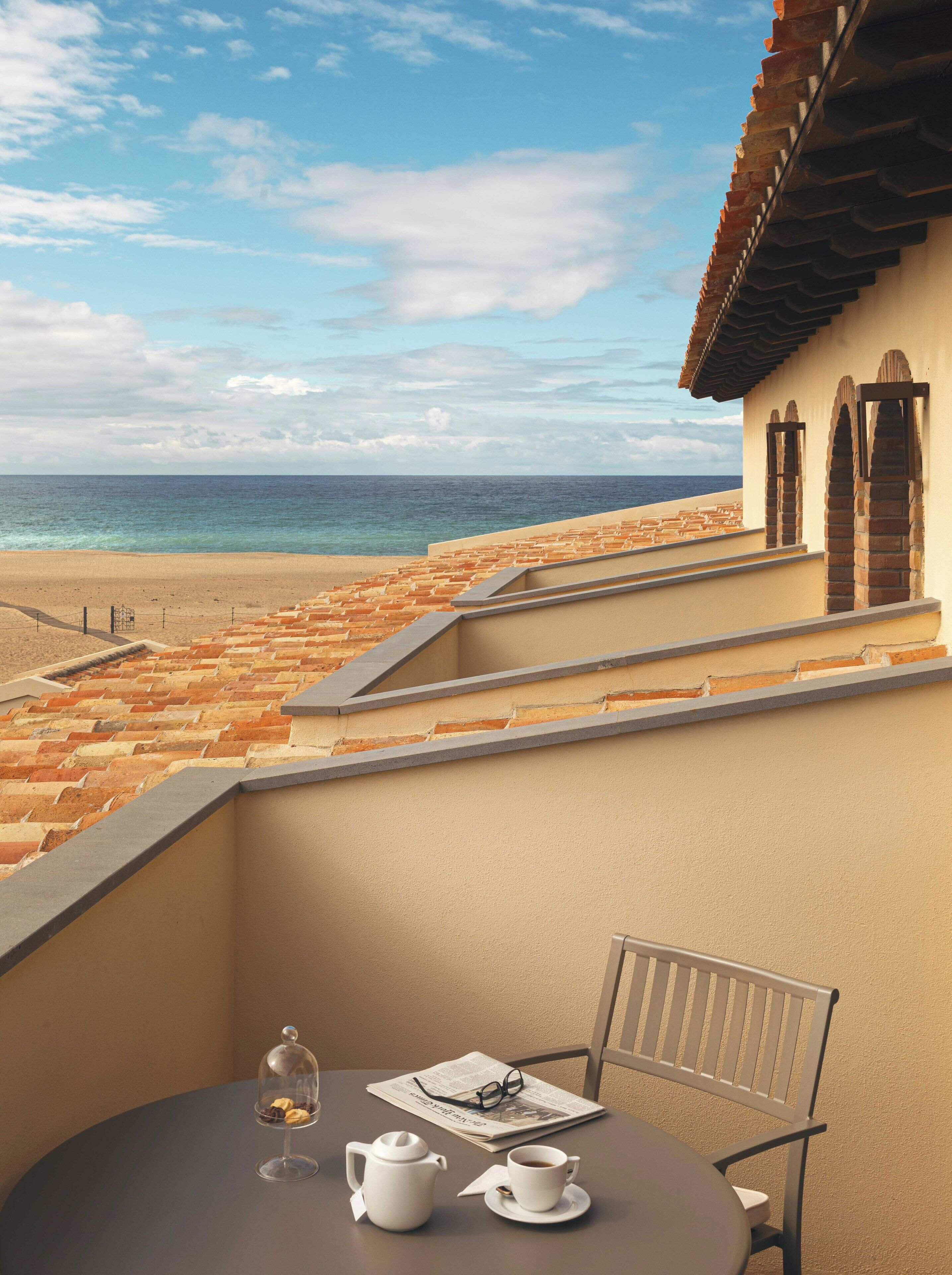 Un balcon avec vue sur la plage et l’océan, une table avec une théière et une tasse, et un journal.