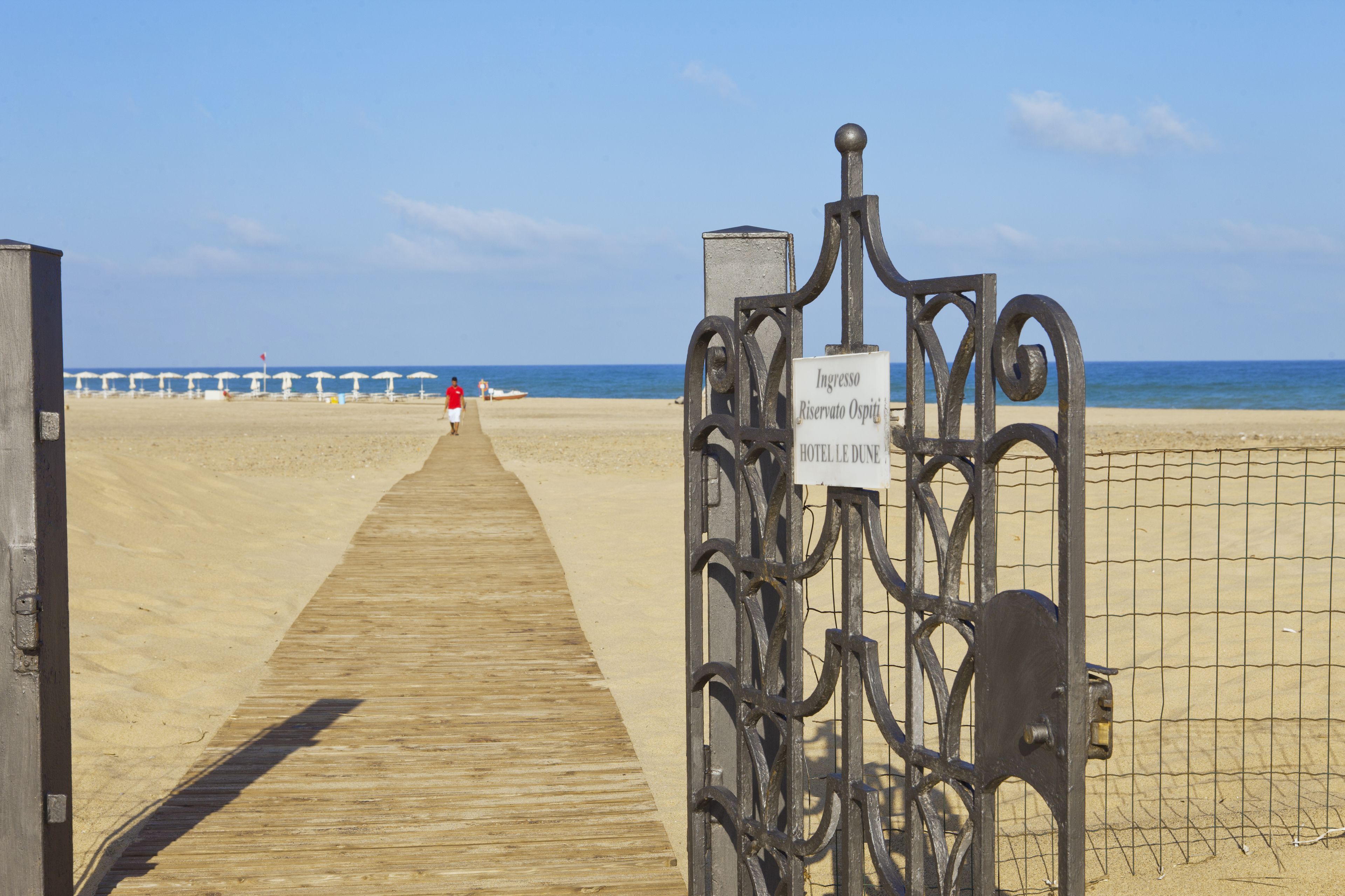 Un muelle de madera que conduce a una playa con una puerta metálica y un letrero que dice 'Ingresso Riservato Ospiti Hotelle Dune'.