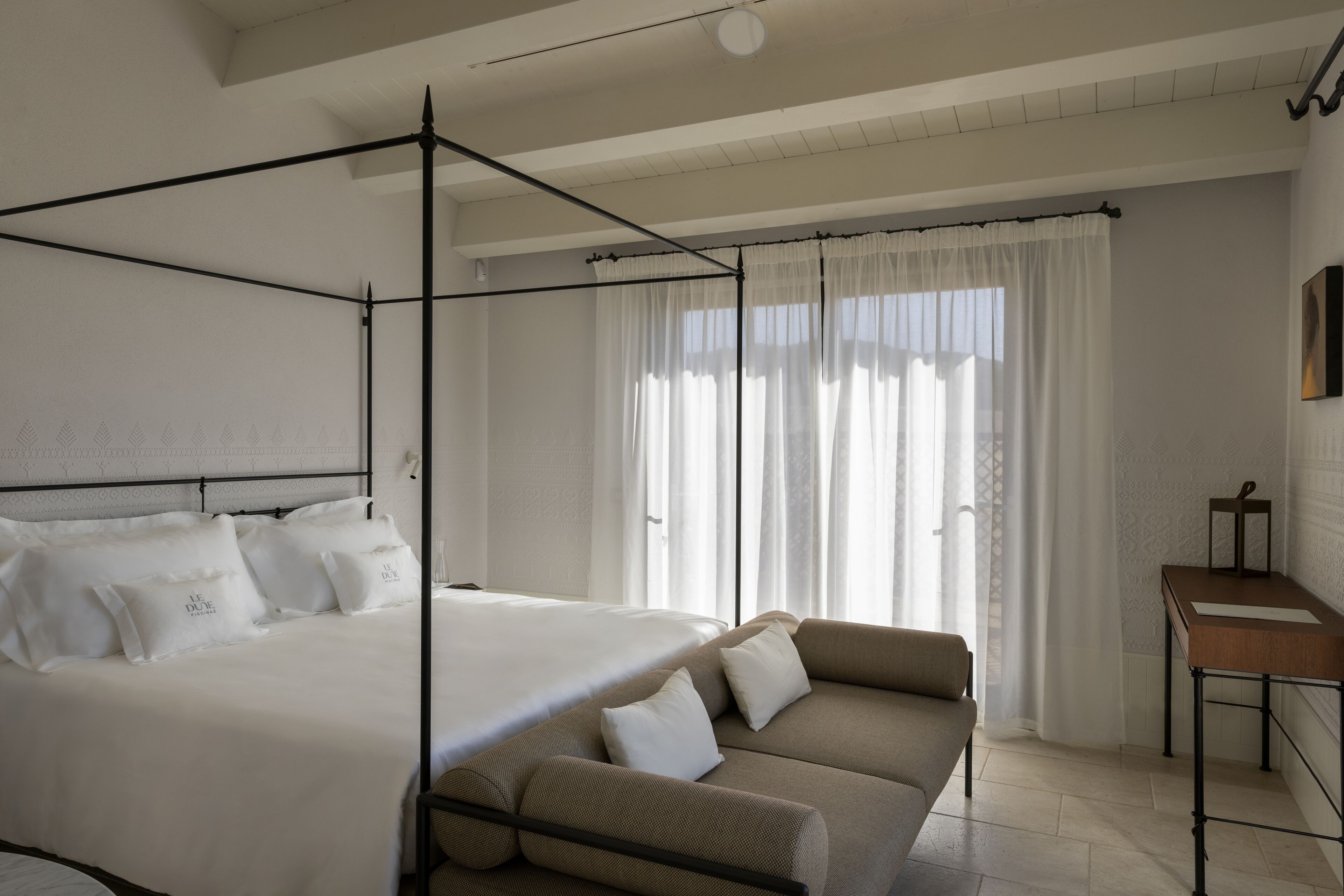 A four-poster bed with white linens and multiple pillows, a beige sofa with cushions, a small wooden side table, and a window with sheer curtains allowing natural light.