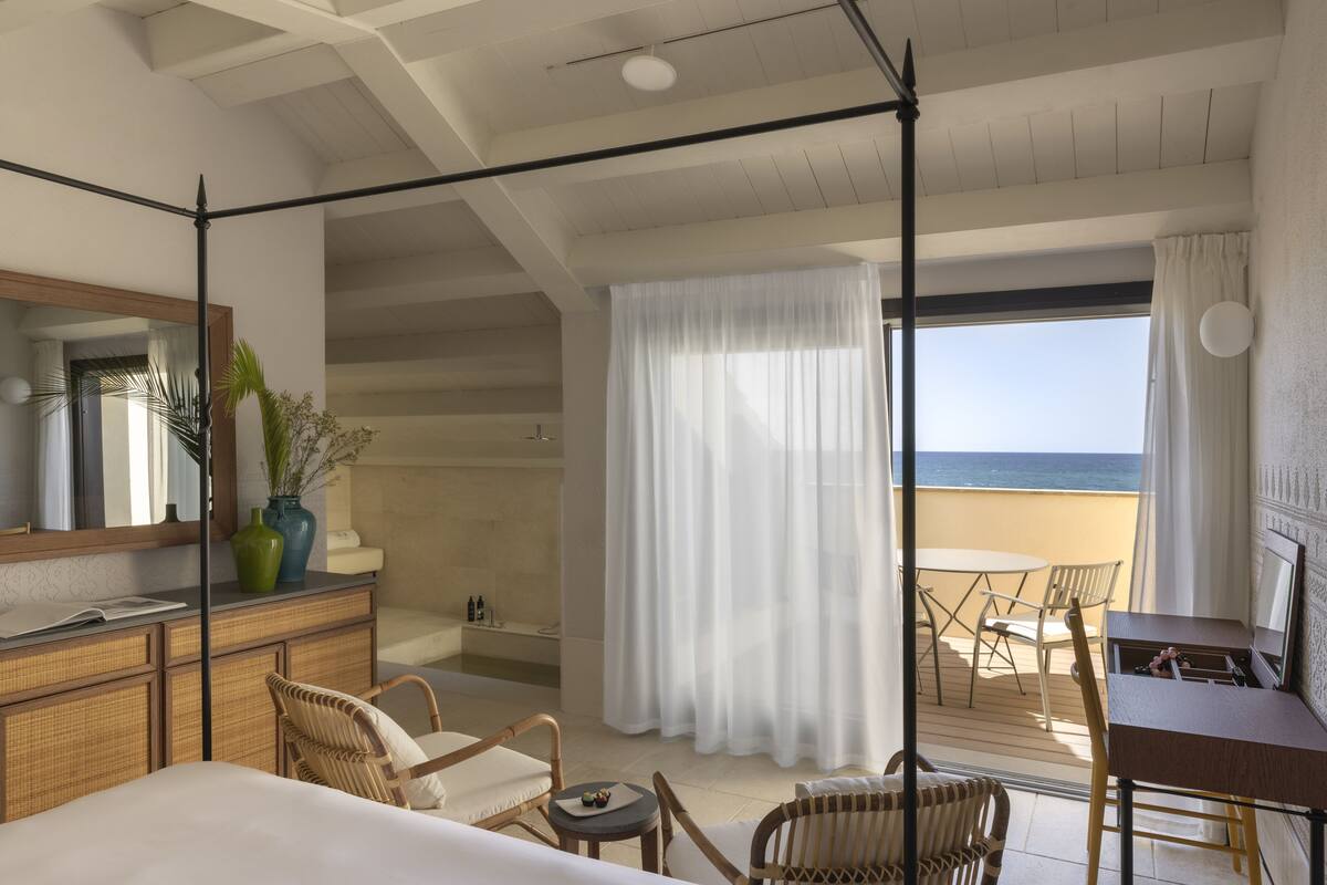 A hotel room with a balcony, a bed, a desk, wicker chairs, a dining table, and a view of the sea.