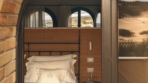 Un dormitorio con pared de ladrillo, una cama con sábanas blancas, una mesita redonda con un vaso, y vista a una playa a través de una ventana.