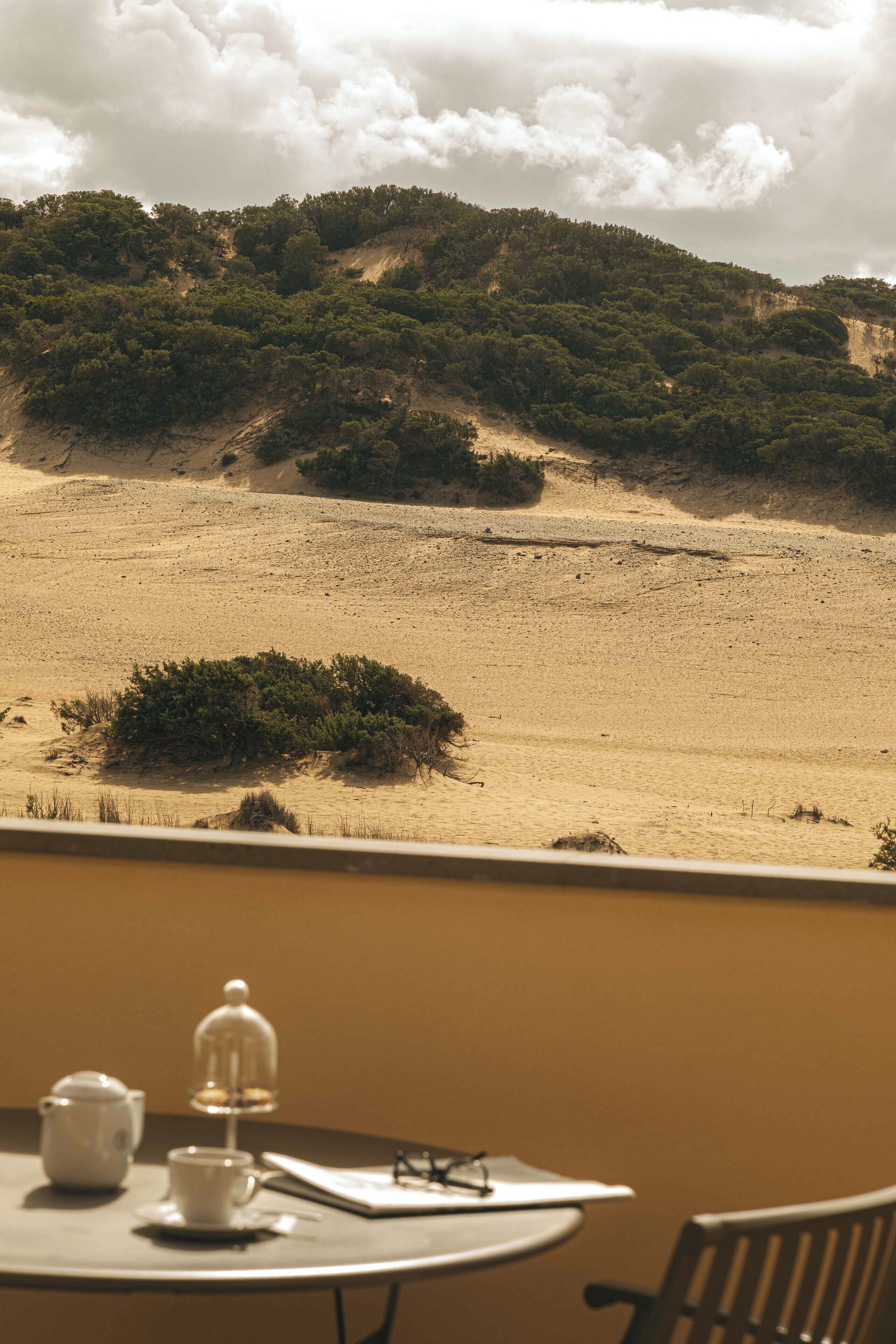 Un tavolo con teiera, tazza e piattino, e una sedia, con vista su un paesaggio desertico con vegetazione rada e colline.