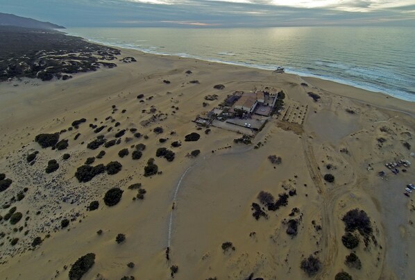 An aerial view of a coastal area with a large sandy beach, scattered vegetation, and a building complex with multiple structures.