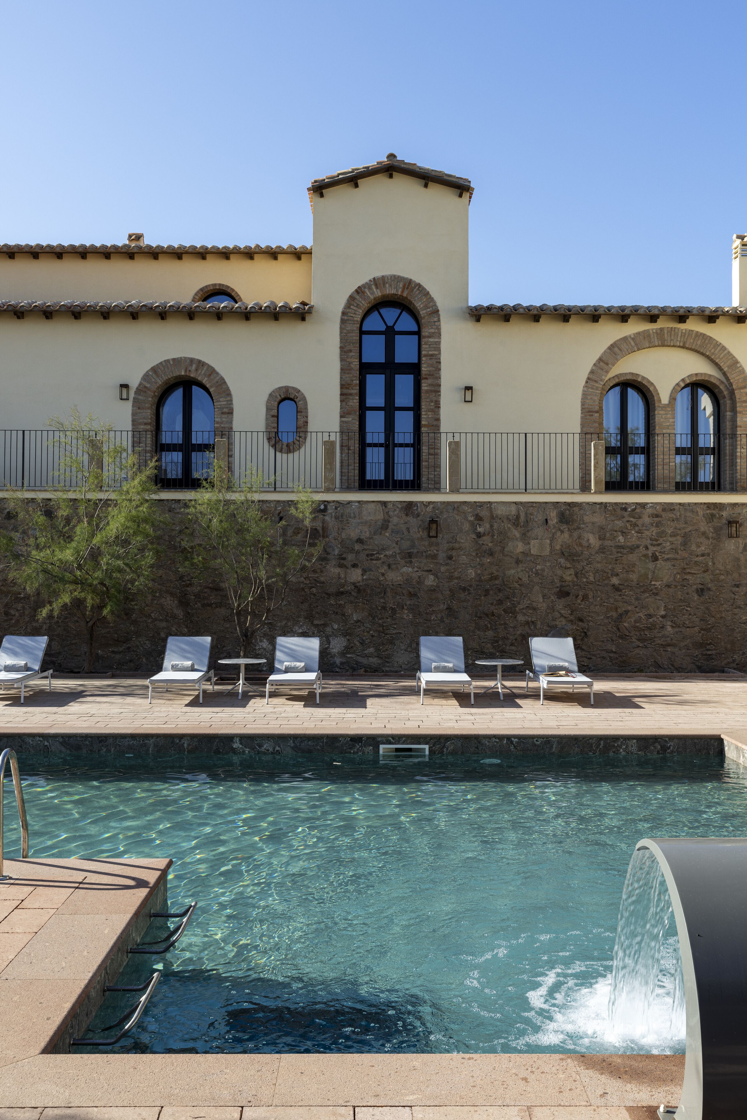 Un área de piscina con agua cristalina, un muro de piedra y un edificio con ventanas arqueadas y un balcón.