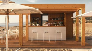 Zona de bar de madera con vistas a la playa, con taburetes y sombrillas.