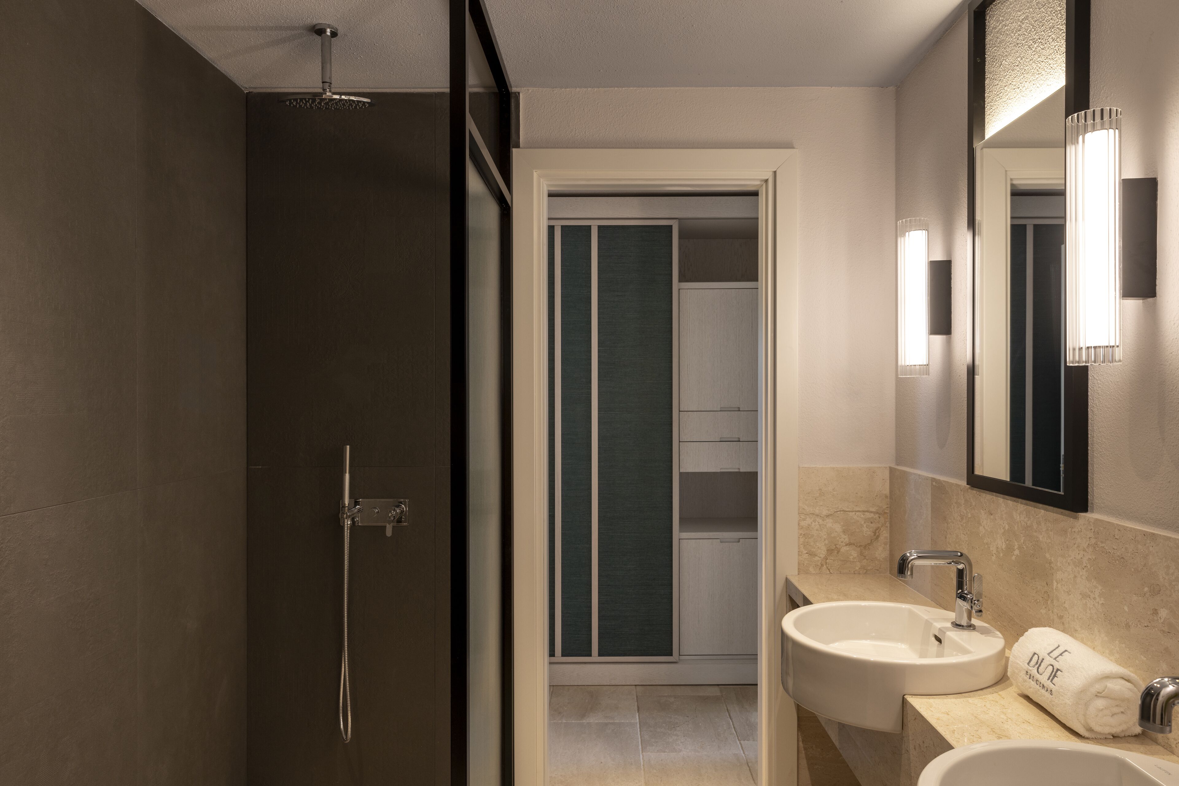 A modern bathroom with a walk-in shower, two sinks, and a mirror with wall-mounted lights.