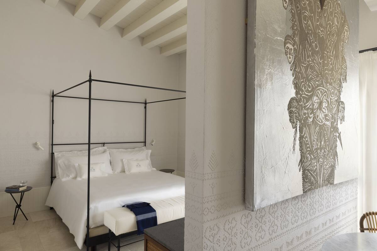 A modern bedroom with a four-poster bed, a small bedside table, and a large decorative wall piece.