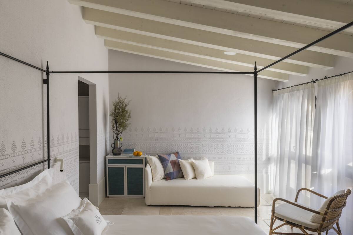 A bedroom with a canopy bed, a bedside table with a vase and fruit, a sofa with patterned cushions, and a wicker chair.