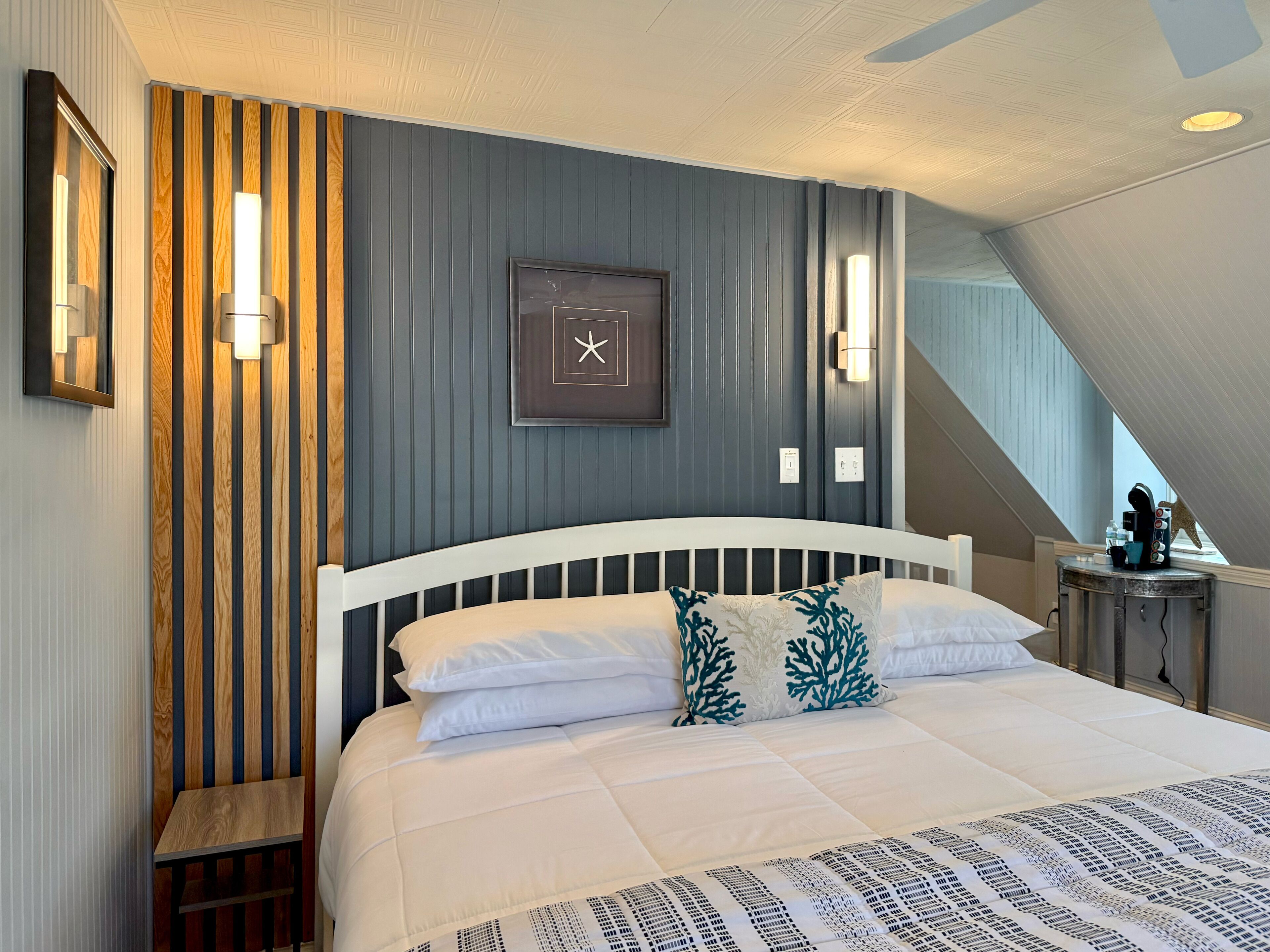 Una habitación de hotel moderna con una cama grande, un panel de madera decorativo, una obra de arte con una estrella de mar enmarcada y una pequeña mesa con cafetera.