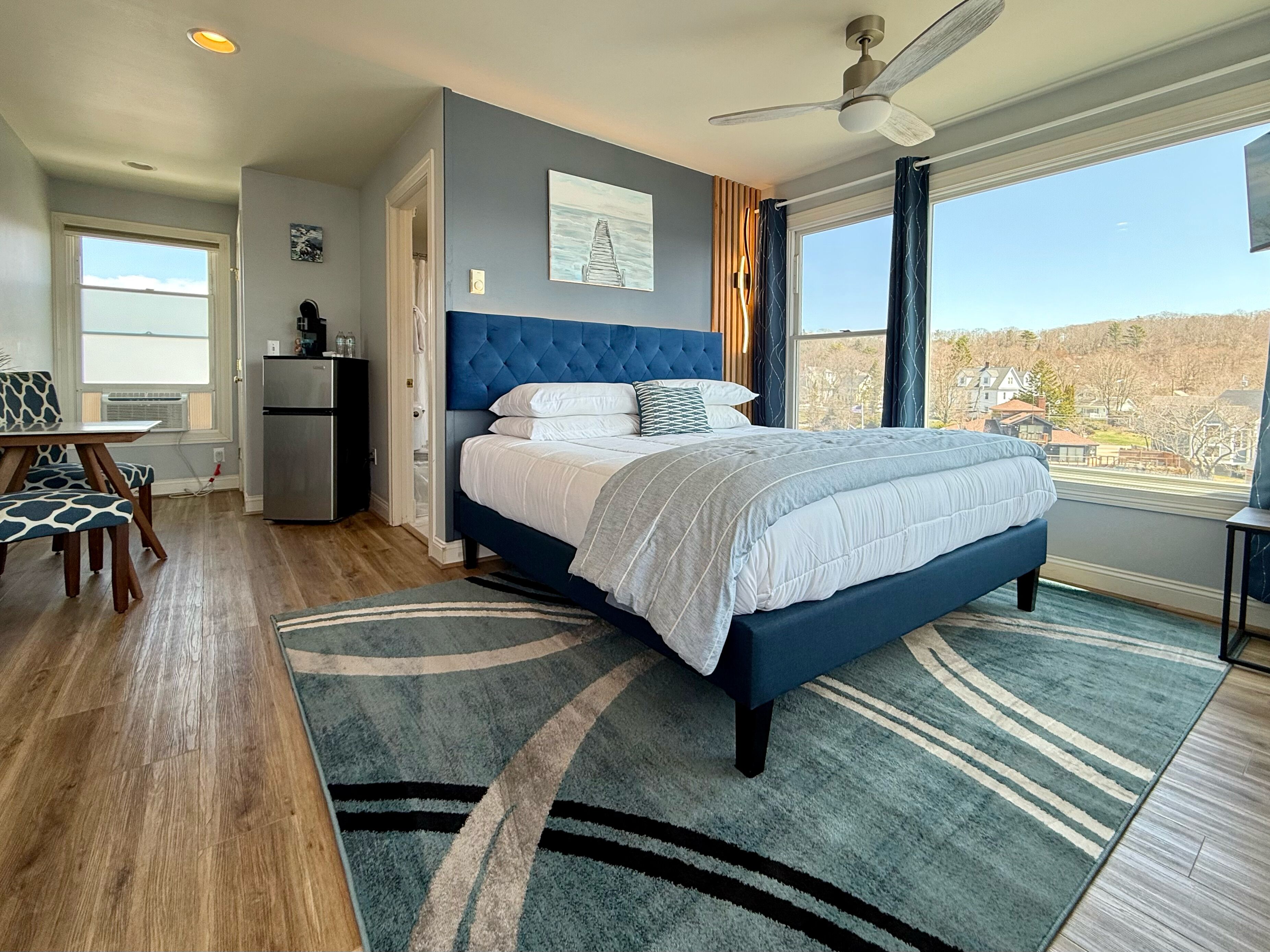 A bedroom with a large bed, a ceiling fan, a view of the outdoors, and a dining area with a table and chairs.