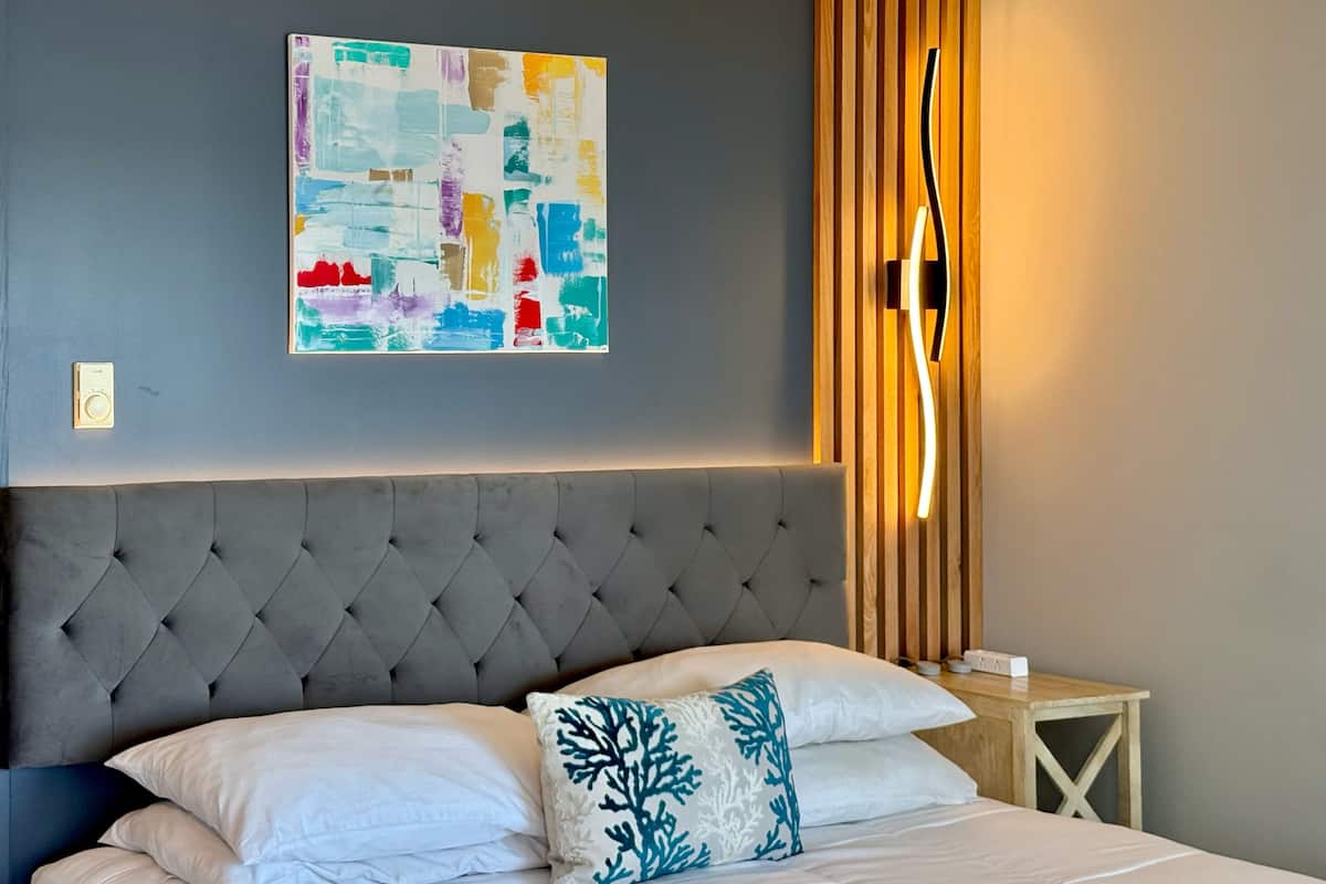 A modern hotel room with a tufted headboard, a large abstract painting, a bedside table with a lamp, and a bed with white linens and decorative pillows.