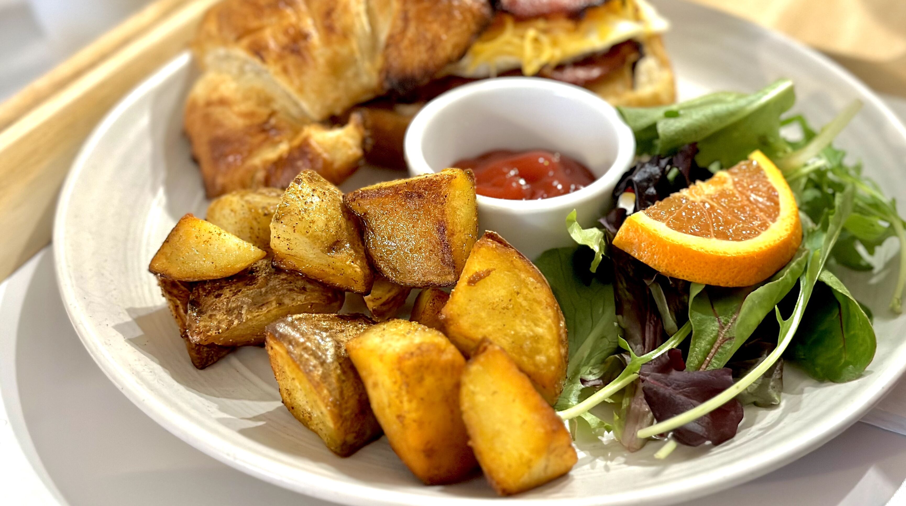 Un plato con papas asadas, un sándwich, una ensalada con una rodaja de naranja y un tazón pequeño de kétchup.