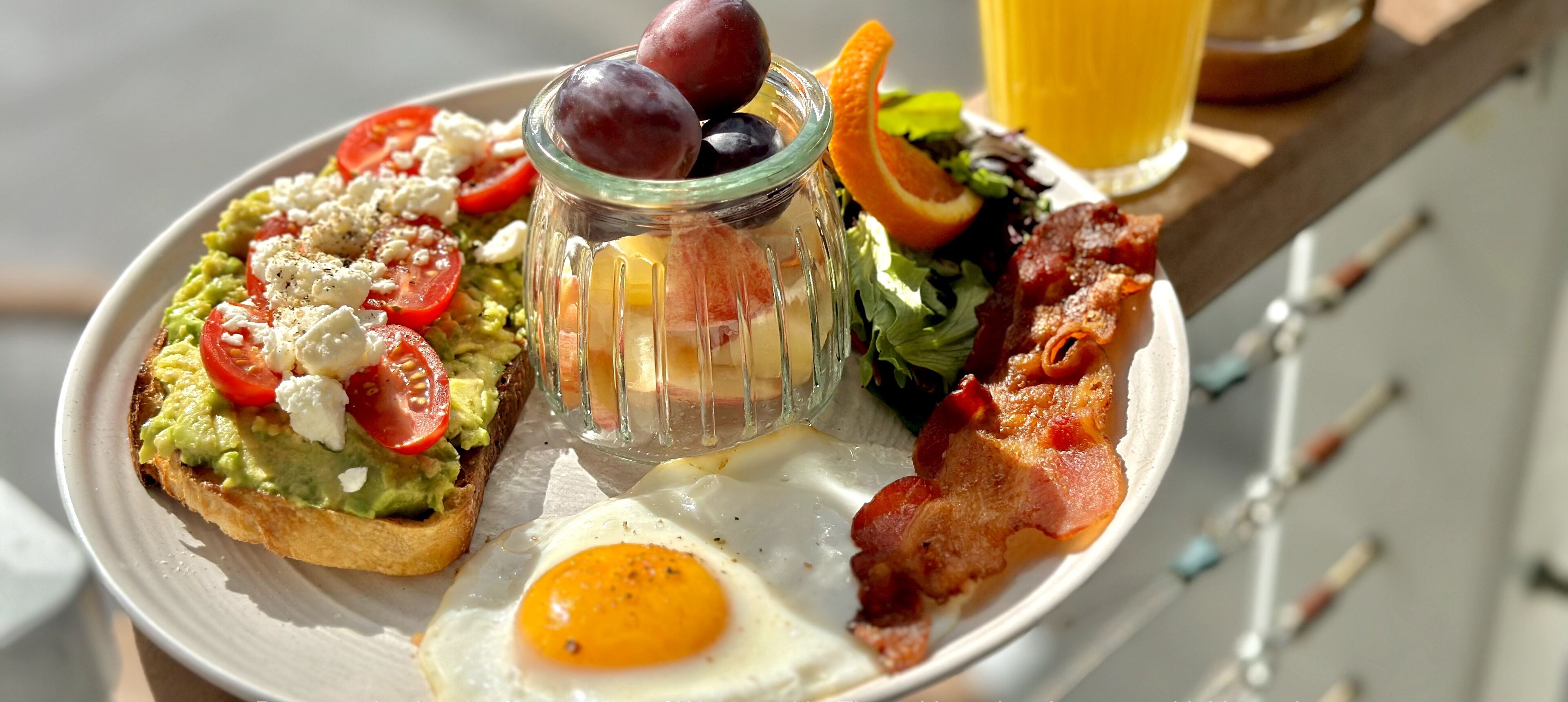 Een ontbijtbord met toast, avocado, tomaten, feta kaas, een gebakken ei, spek, een salade en een glas sinaasappelsap.