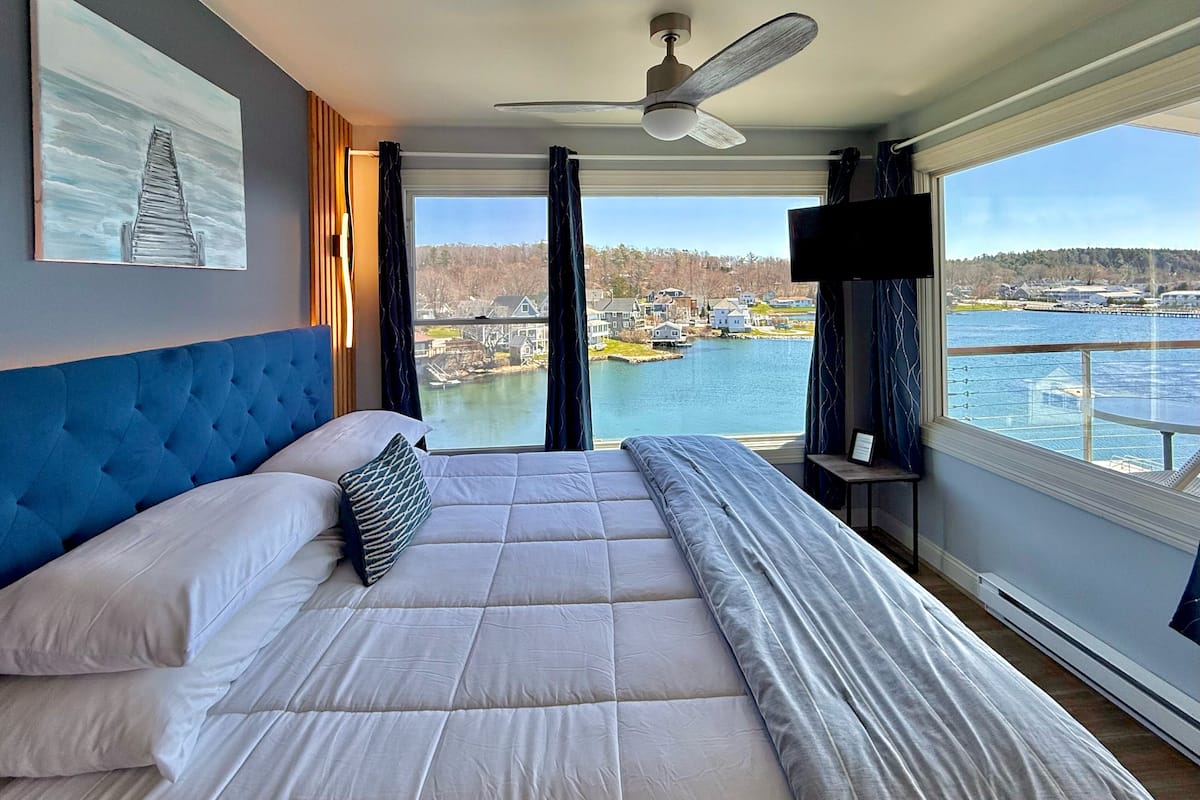 A bedroom with a large bed, a ceiling fan, a TV, and a view of a body of water and buildings.