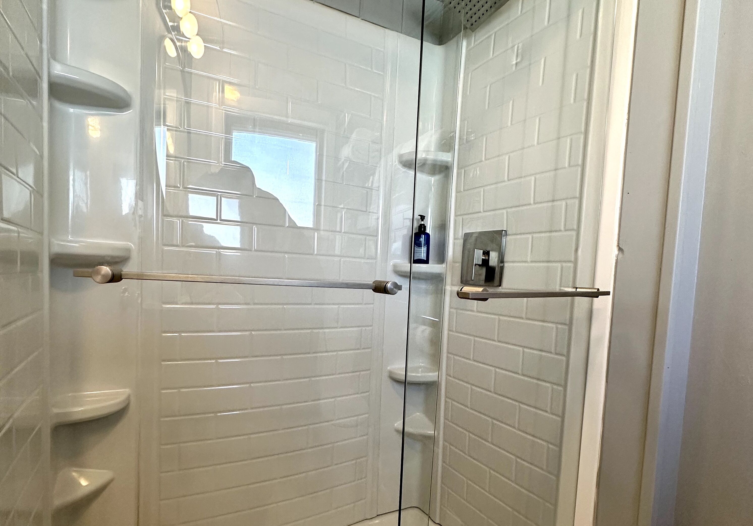 Un baño moderno con azulejos blancos tipo metro, cerramiento de ducha de vidrio y una botella de champú a la vista.