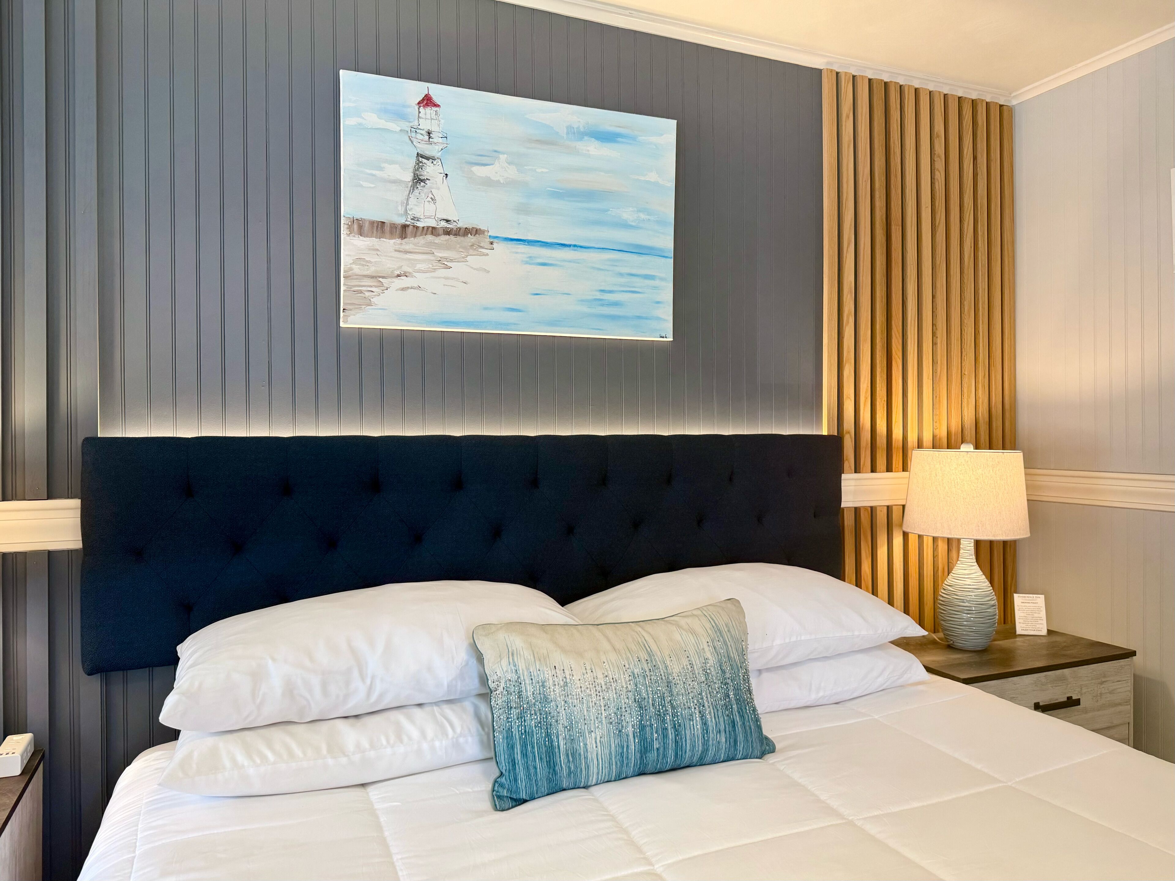 Une chambre d’hôtel avec un grand lit, une tête de lit ornée de panneaux verticaux, un tableau représentant un phare, une table de chevet avec une lampe et un mur d’accent en bois.