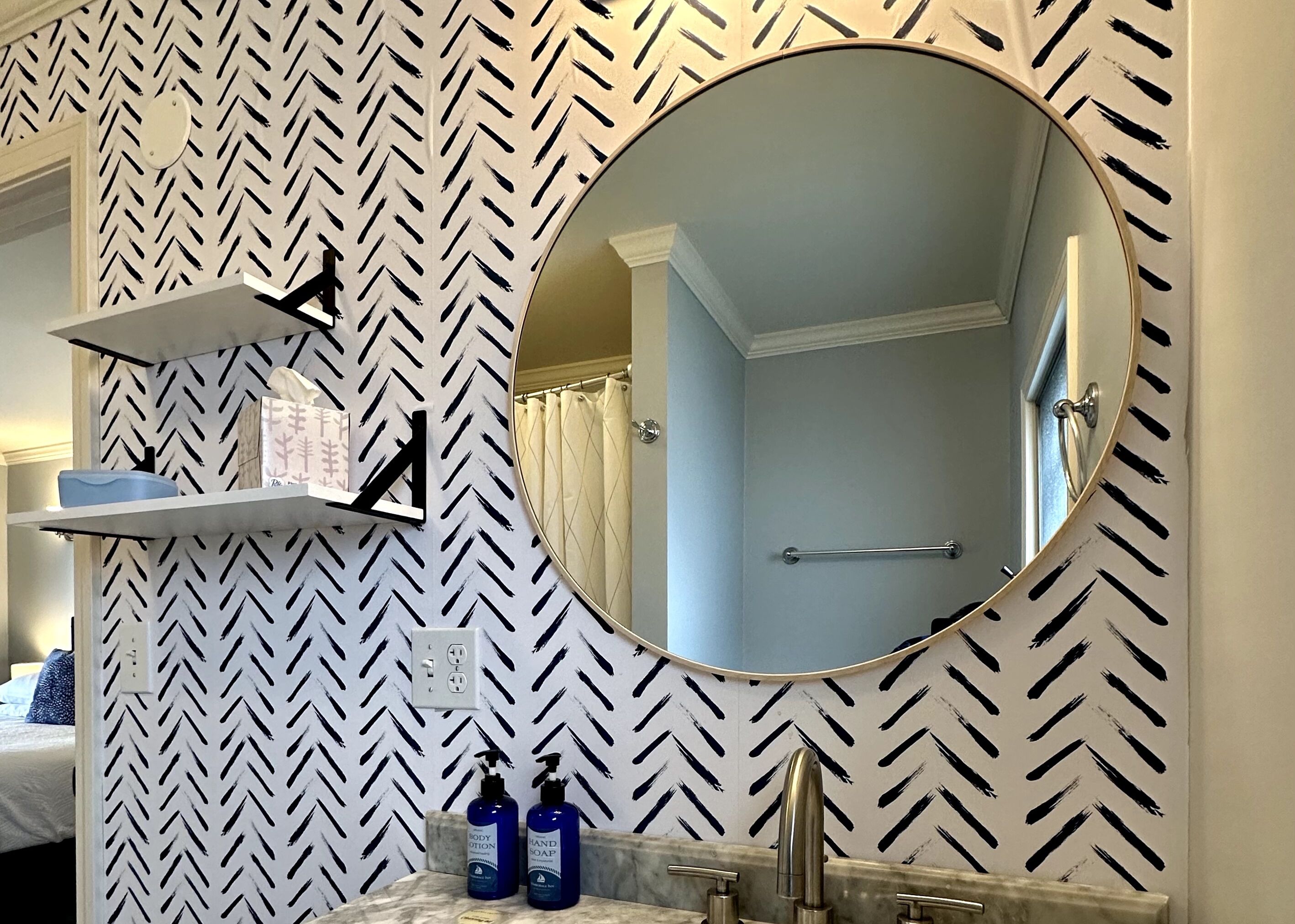 A bathroom with a mirror, a towel rack, a shelf with toiletries, and a patterned wall.