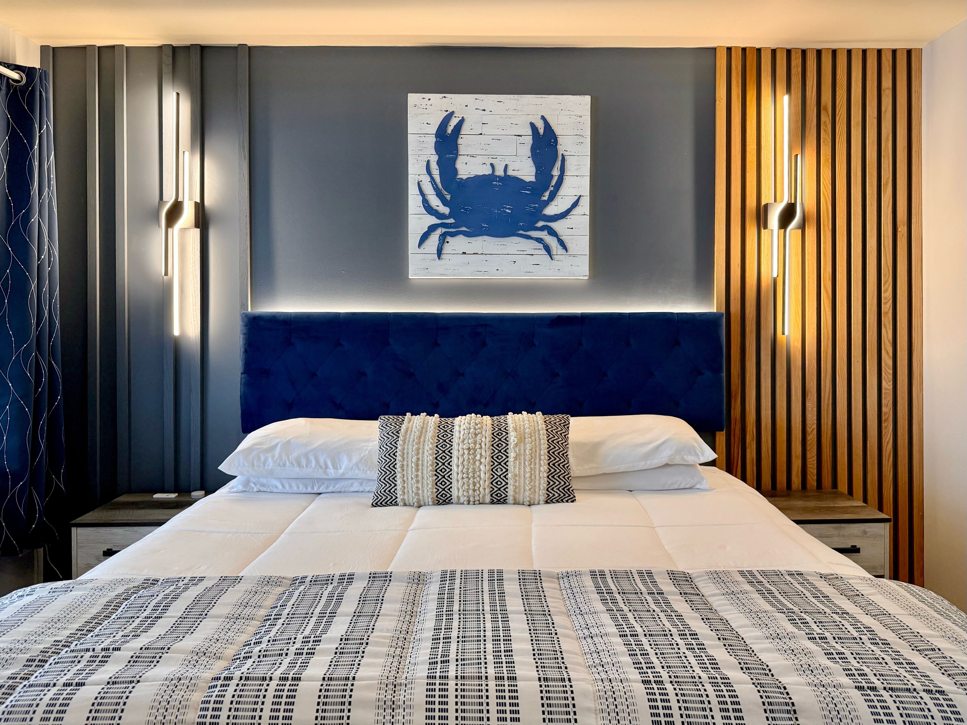 A modern hotel room with a large bed, a blue headboard, a wall-mounted artwork of a crab, and vertical wooden slats on one wall.