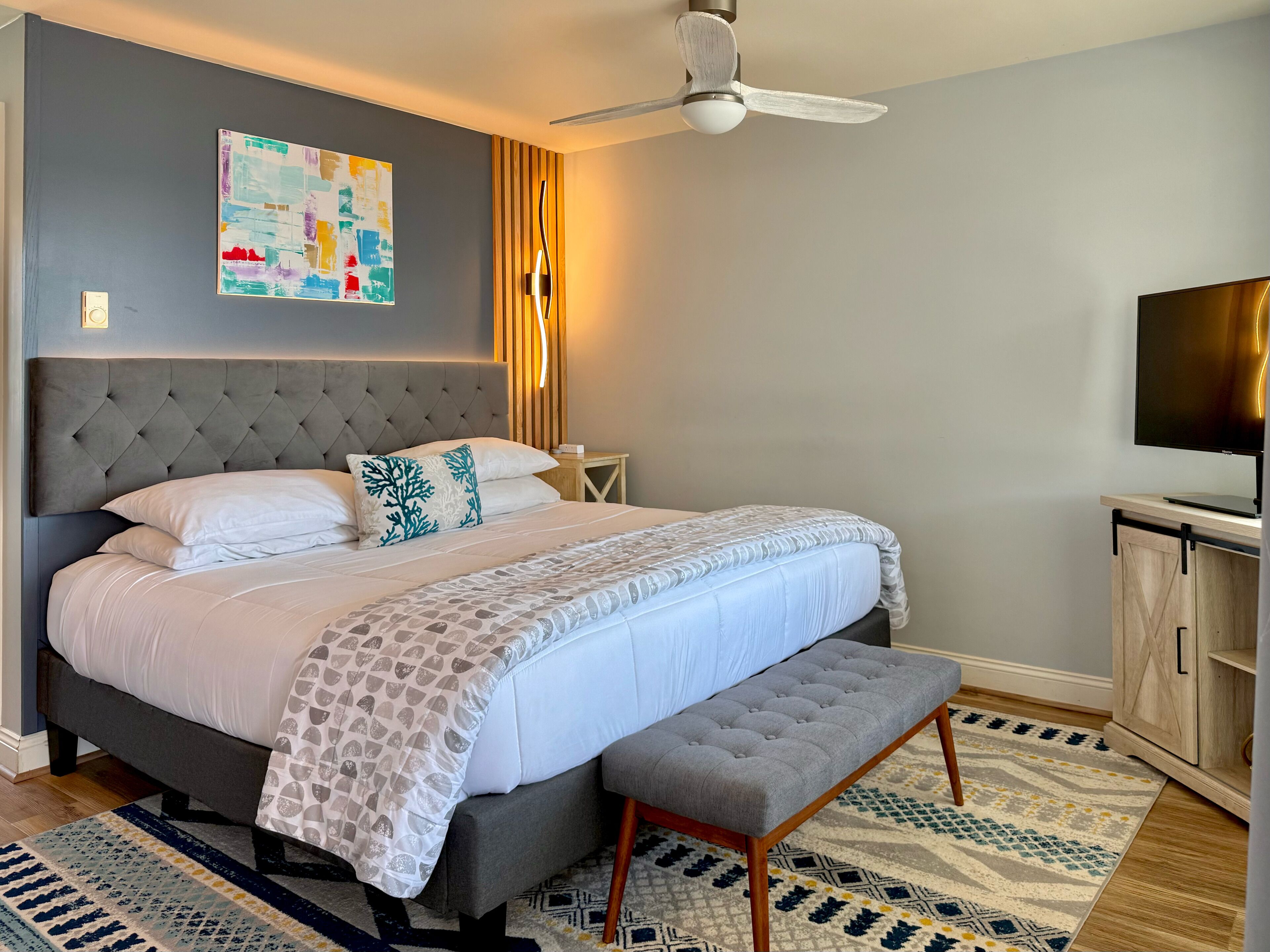 A modern bedroom with a tufted headboard, a large bed with white bedding, a bench, a ceiling fan, a wall-mounted TV, and a patterned rug.