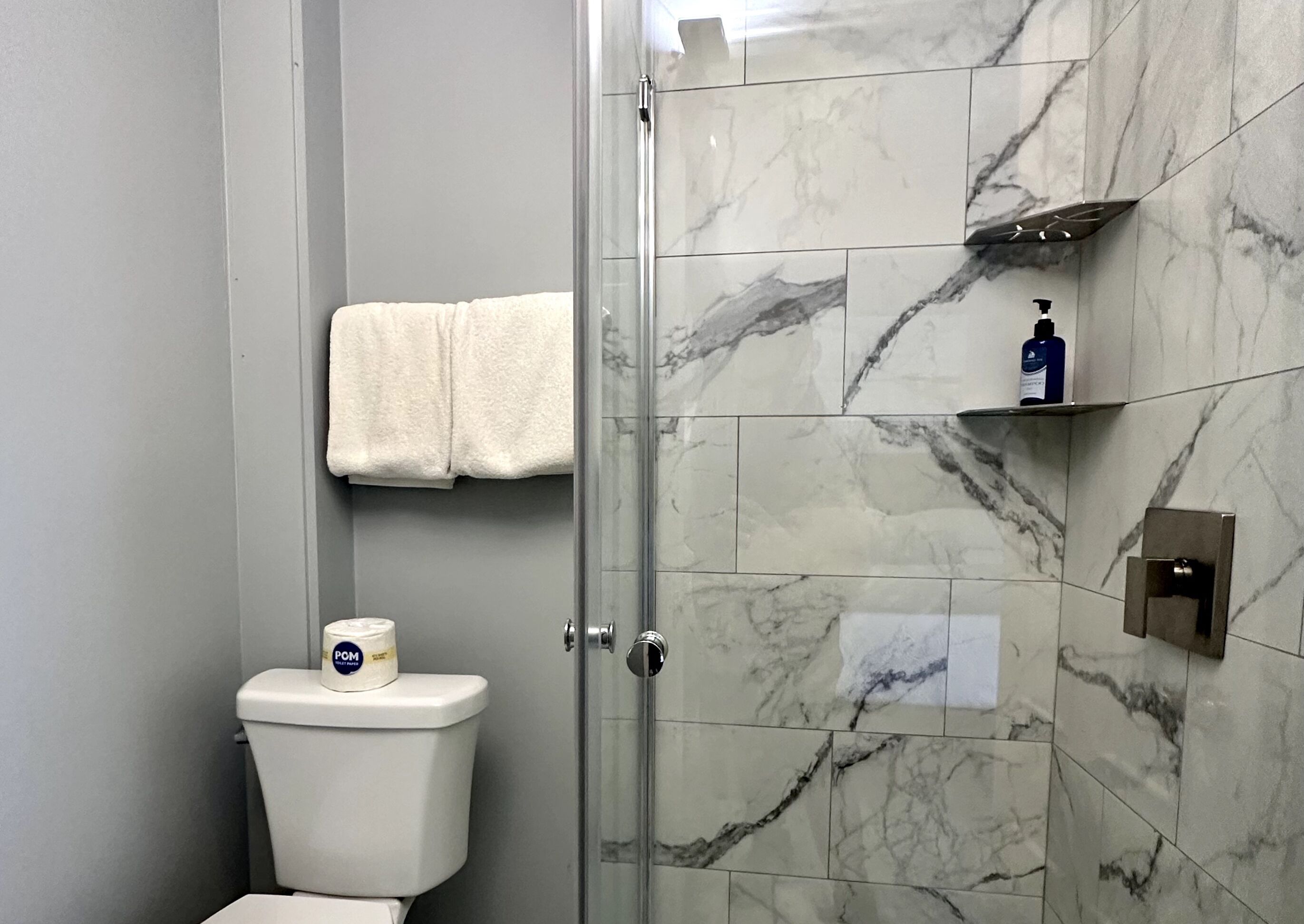 Et badeværelse med toilet, bruseniche, marmorfliser, et håndklæde på væggen og en sæbedispenser på hylden.