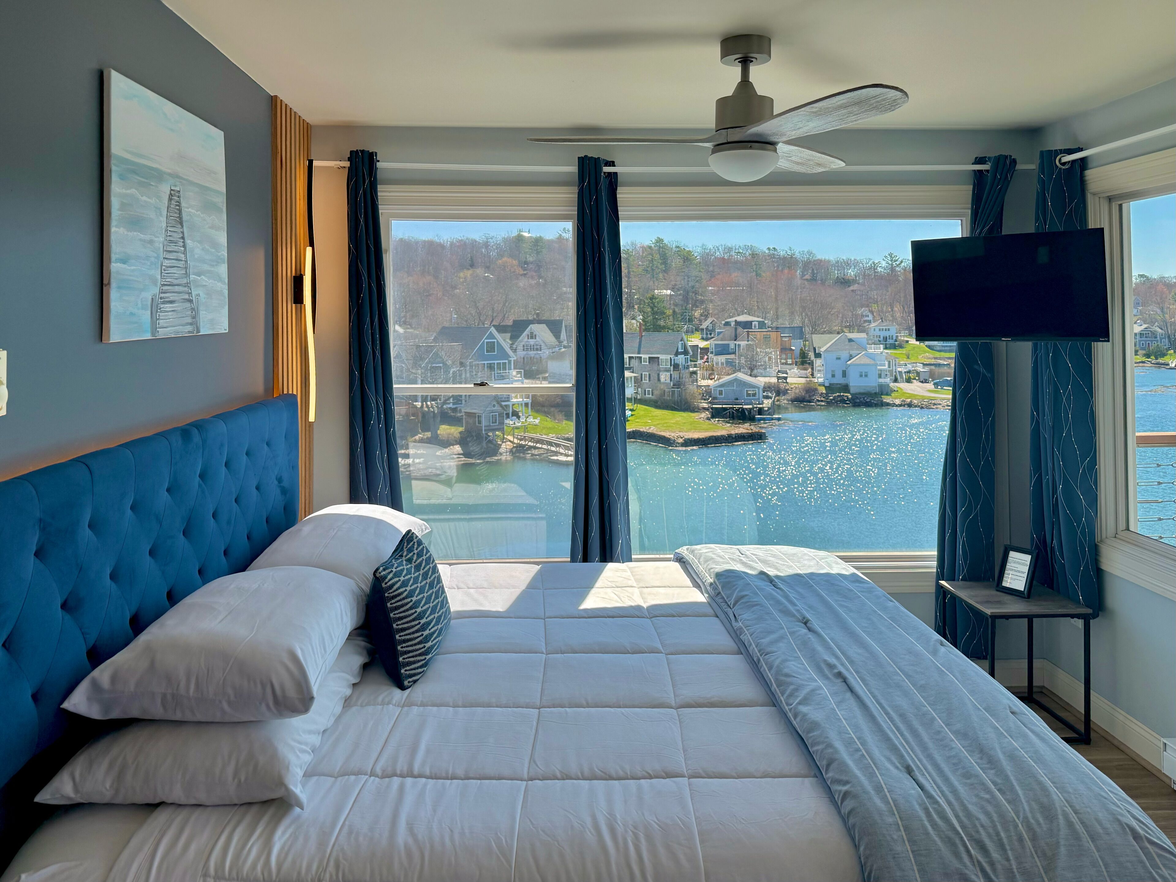 Une chambre avec un grand lit, un ventilateur de plafond, une télévision et une vue sur un lac et des maisons.