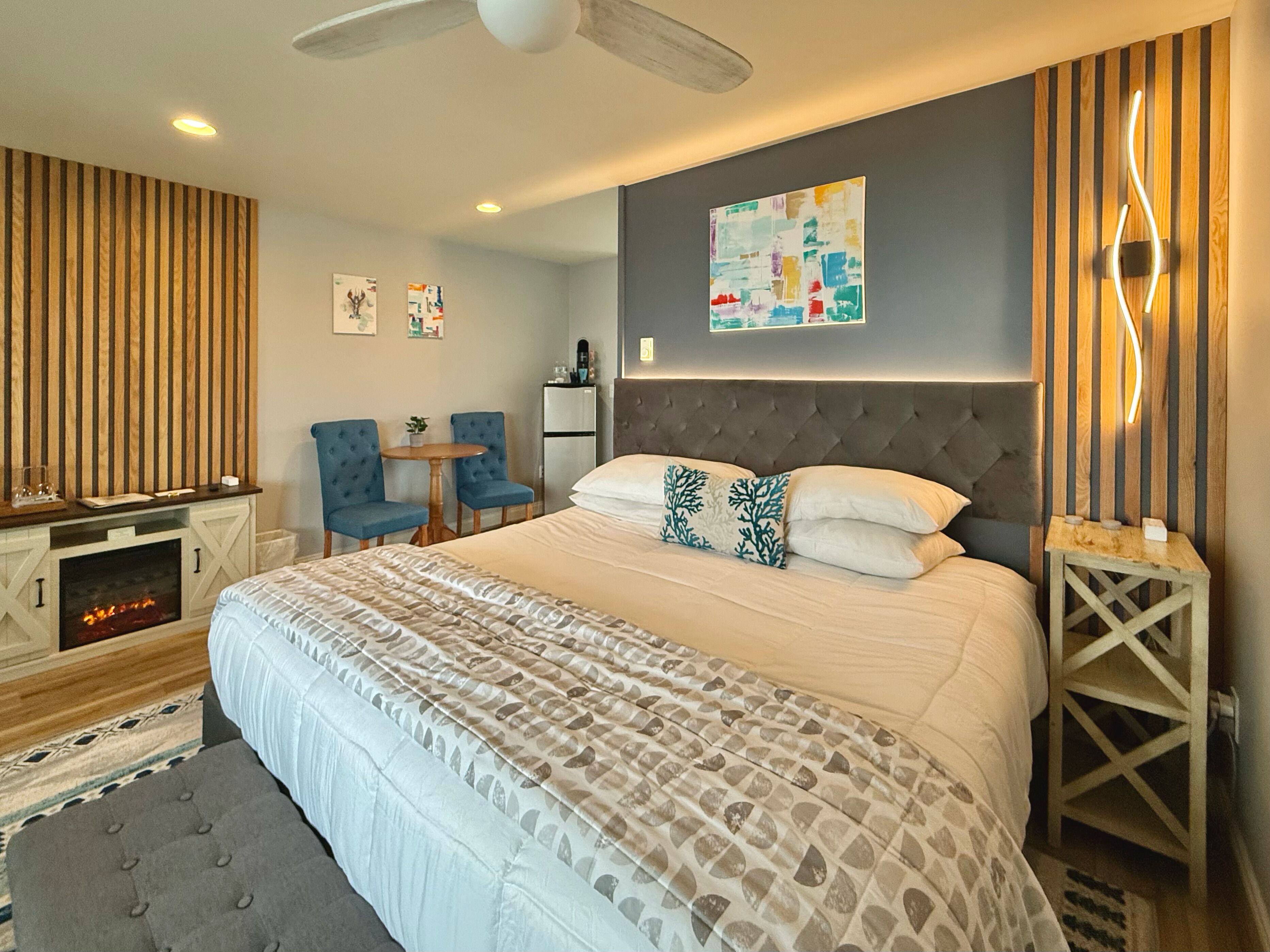 Une chambre moderne avec un grand lit, une cheminée, deux fauteuils, une petite table et une œuvre d’art accrochée au mur.