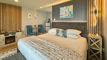 A modern bedroom with a large bed, a fireplace, two chairs, a small table, and a wall-mounted artwork.
