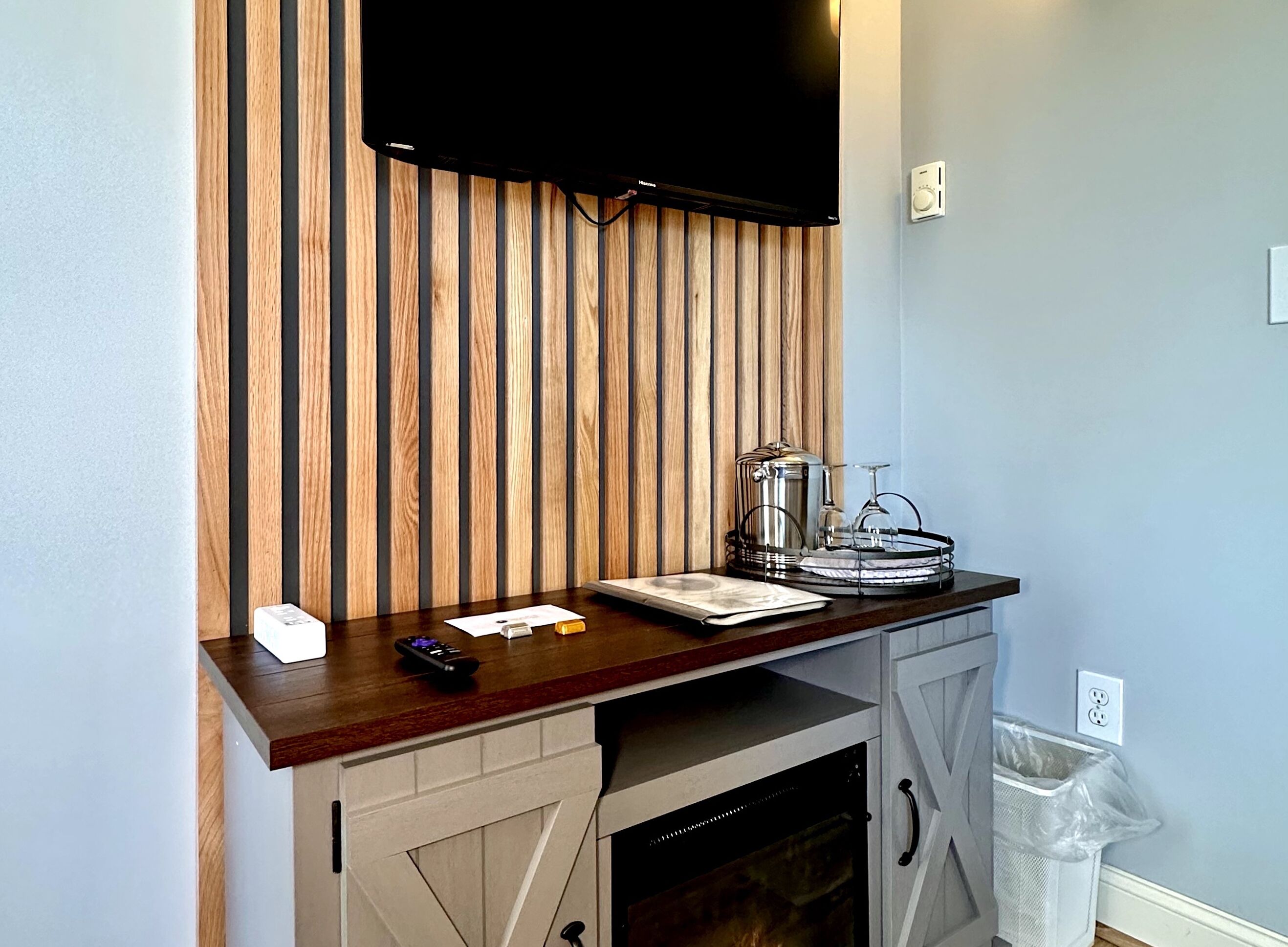 A room with a wooden slat wall, a flat-screen TV mounted above a wooden counter, a trash bin, and a power outlet on the wall.