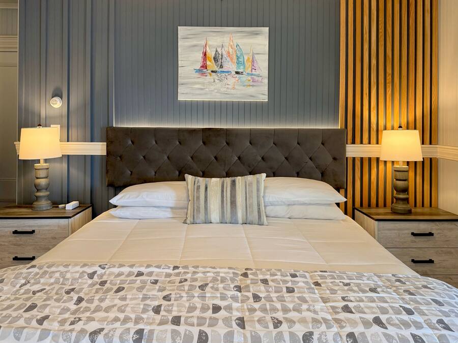 Una habitación de hotel con una cama grande, dos mesitas de noche con lámparas, un cabecero capitonado y un cuadro de barcos a vela en la pared.