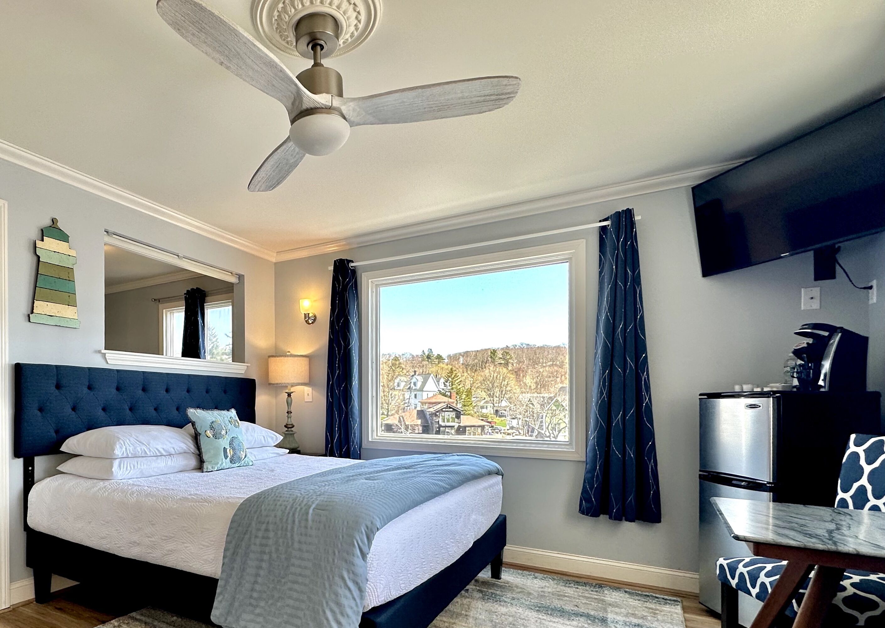 Une chambre avec un lit, un ventilateur de plafond, une télévision, une fenêtre avec des rideaux et un coin cuisine.