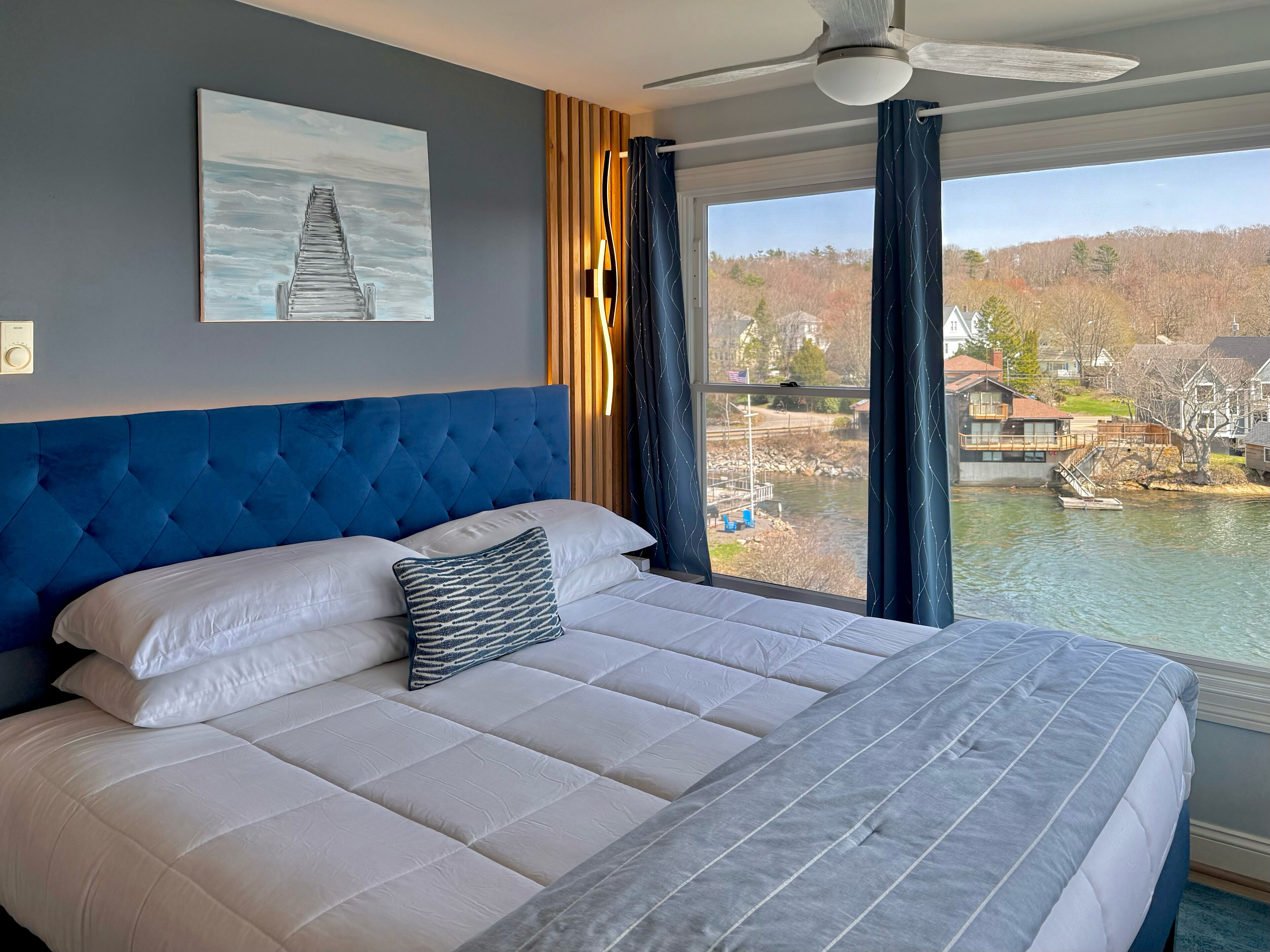 A bedroom with a large bed, a ceiling fan, a painting on the wall, and a view of a lake and buildings.