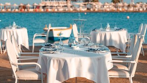Ein Restaurant direkt am Strand mit runden Tischen und weißen Stühlen, mit Blick auf das Meer.