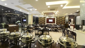 Un grand espace dédié à la salle à manger, avec de nombreuses tables et chaises, un buffet disposant de présentoirs couverts pour les plats, et un grand écran diffusant du contenu.