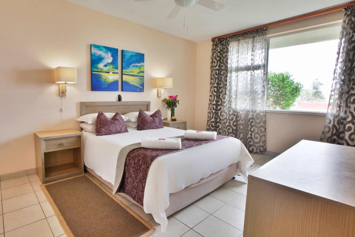Habitación de hotel con cama, lámparas de noche, mesita de noche, escritorio, ventana con cortinas y obra de arte en la pared.
