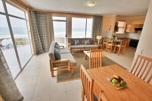 Amplia sala de estar con sofá, mesa de comedor y sillas, cocina con armarios de madera y ventanas grandes con vista al océano.
