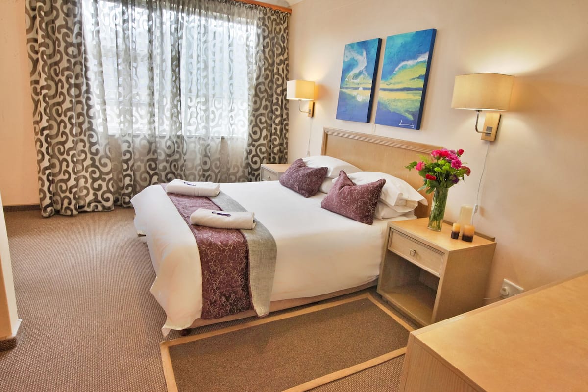 Habitación de hotel con una cama grande, dos lámparas fijadas a la pared, una mesita de noche con un florero y un cuadro sobre la cama.