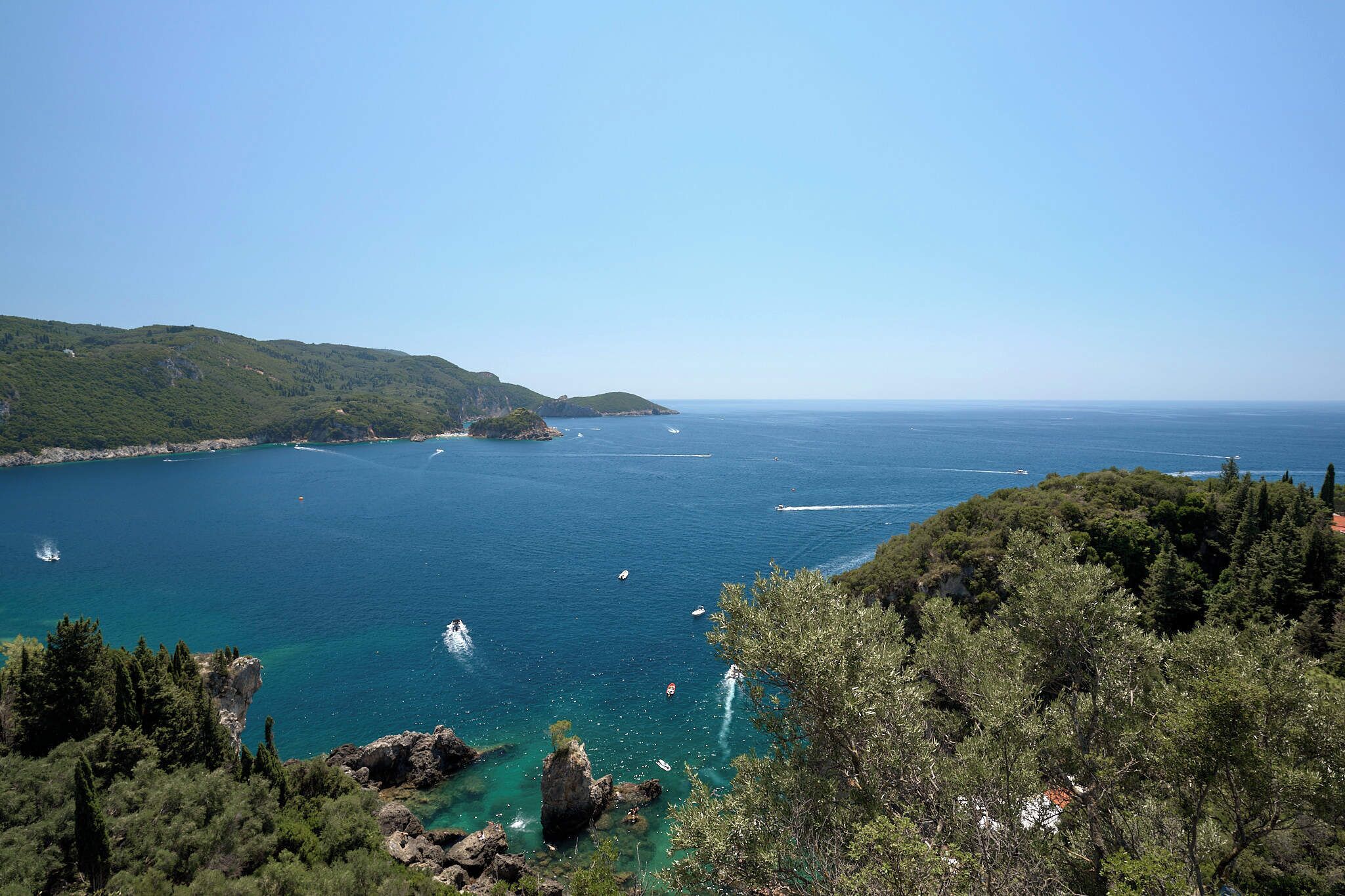 A coastal landscape with clear blue sea, rocky outcrops, and dense greenery.
