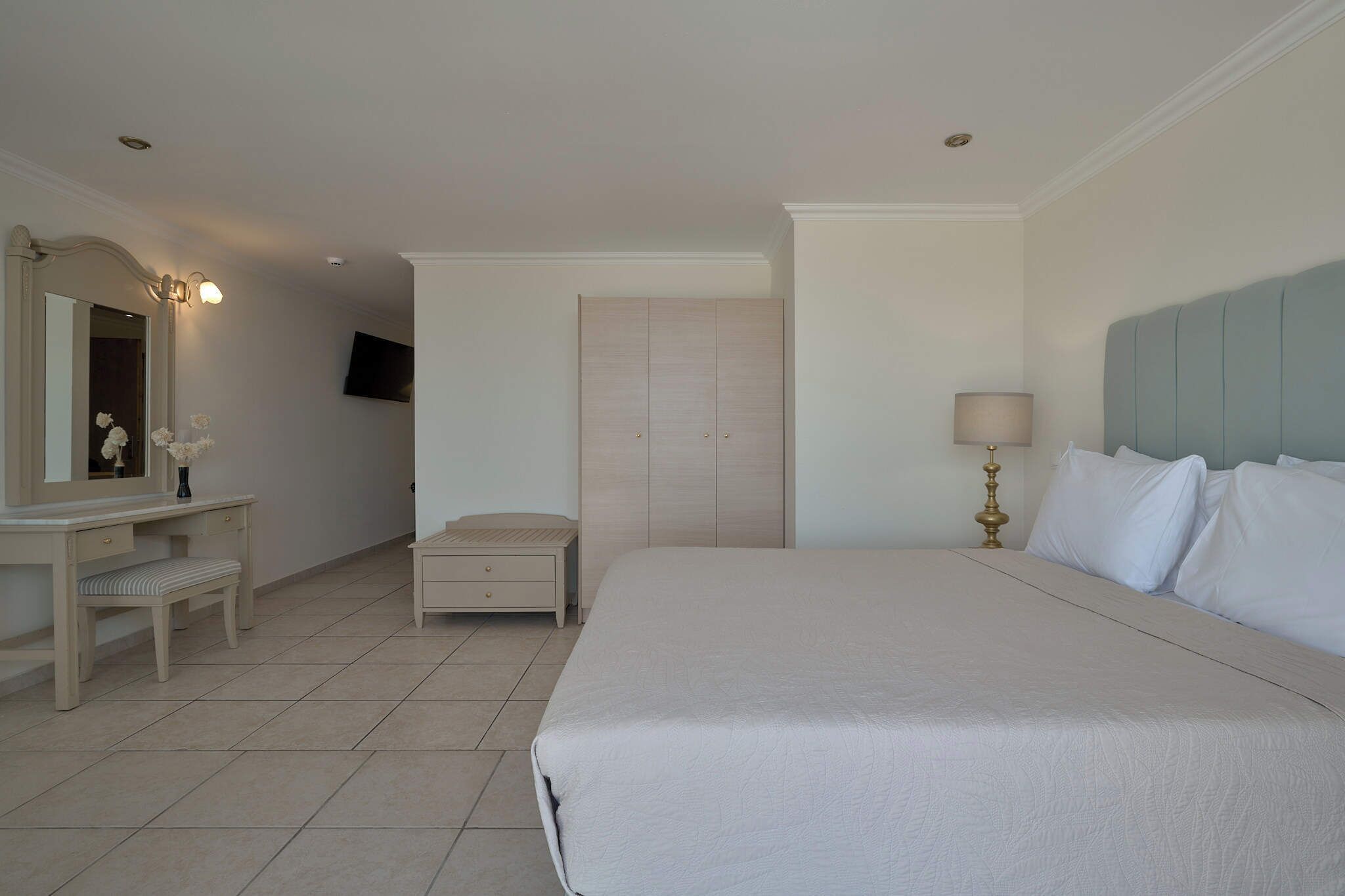 Quarto de hotel com cama, mesa de cabeceira, espelho, cômoda, televisão e abajur.