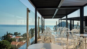A terrace with clear glass walls, tables, and chairs, overlooking a coastal view. - Paleokastritsa Palace (Corfu)
