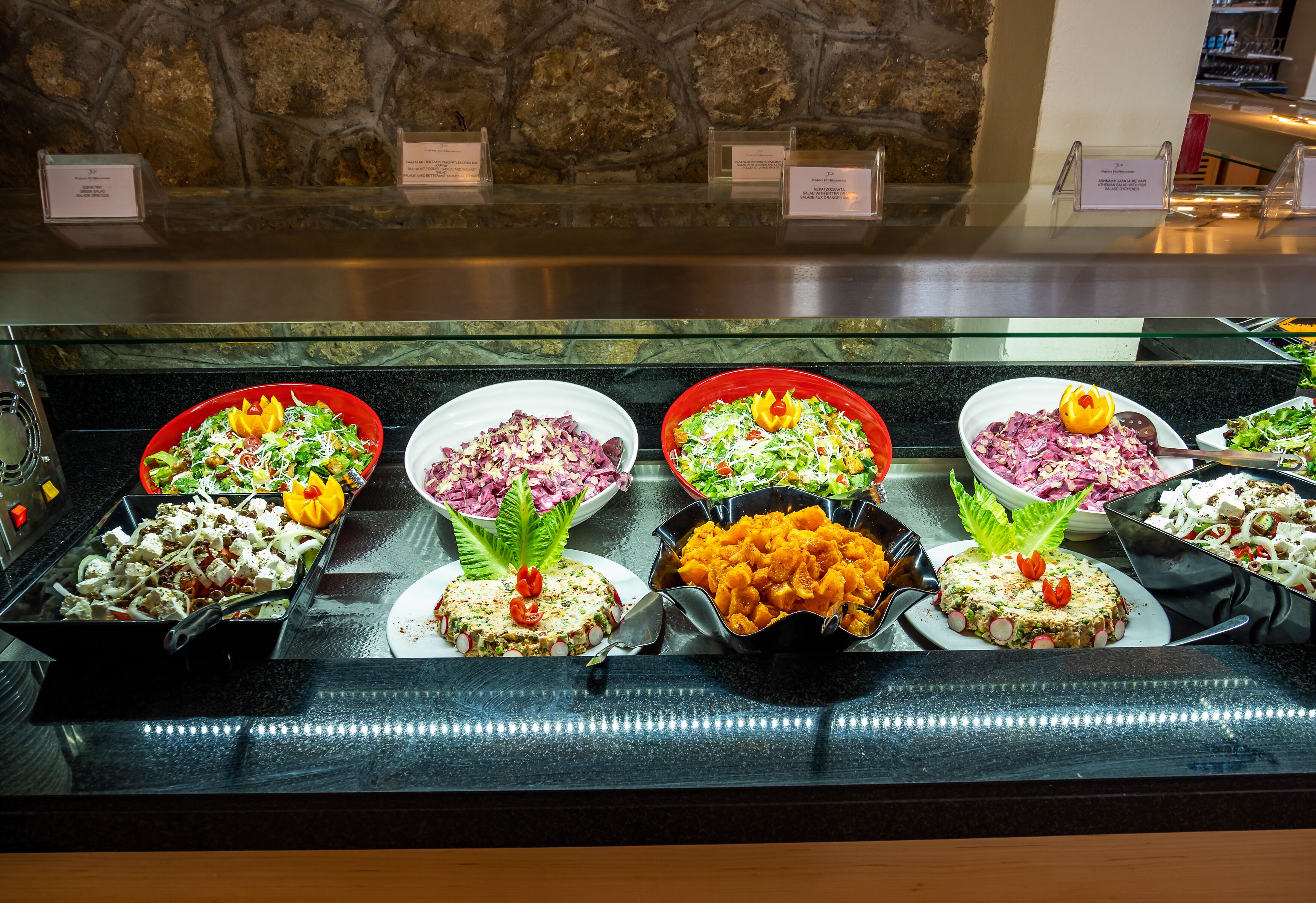 A buffet with various dishes, including salads and a meat-based dish, displayed in a glass case with a stone wall background.